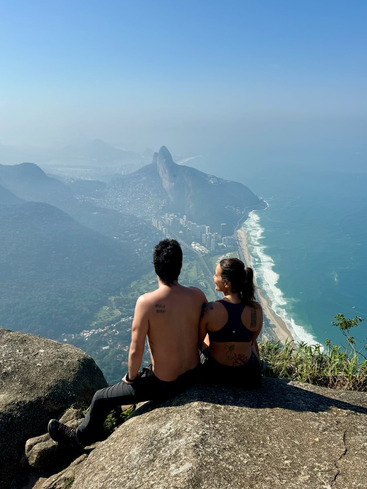 Thaíssa e Julián sentados numa pedra com a cidade e o mar do Rio ao fundo
