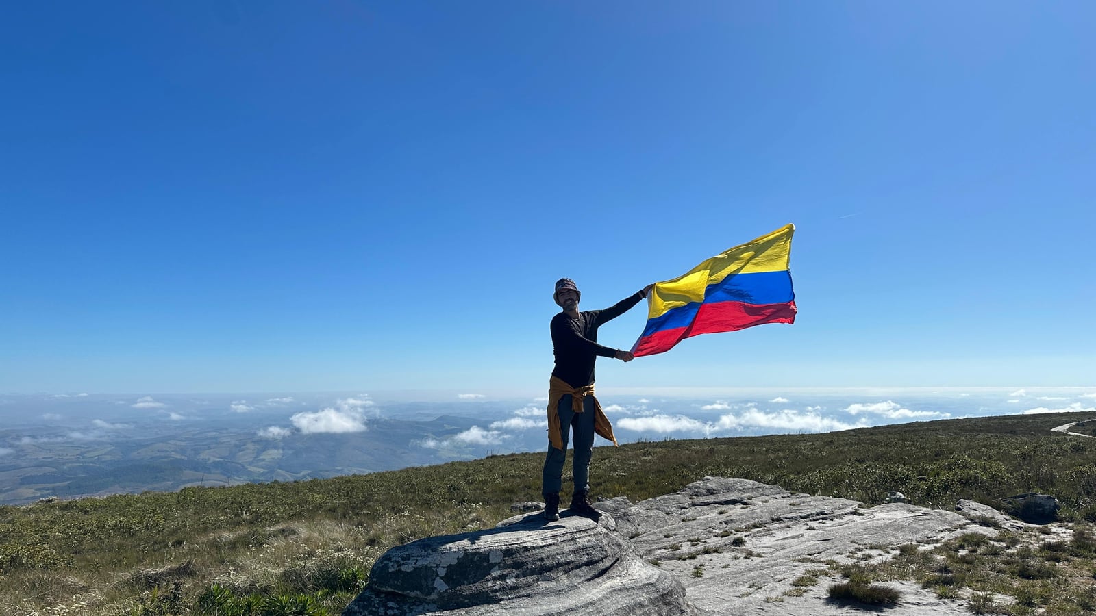 Julián de pé numa pedra acima das nuvens, segurando a bandeira da Colômbia
