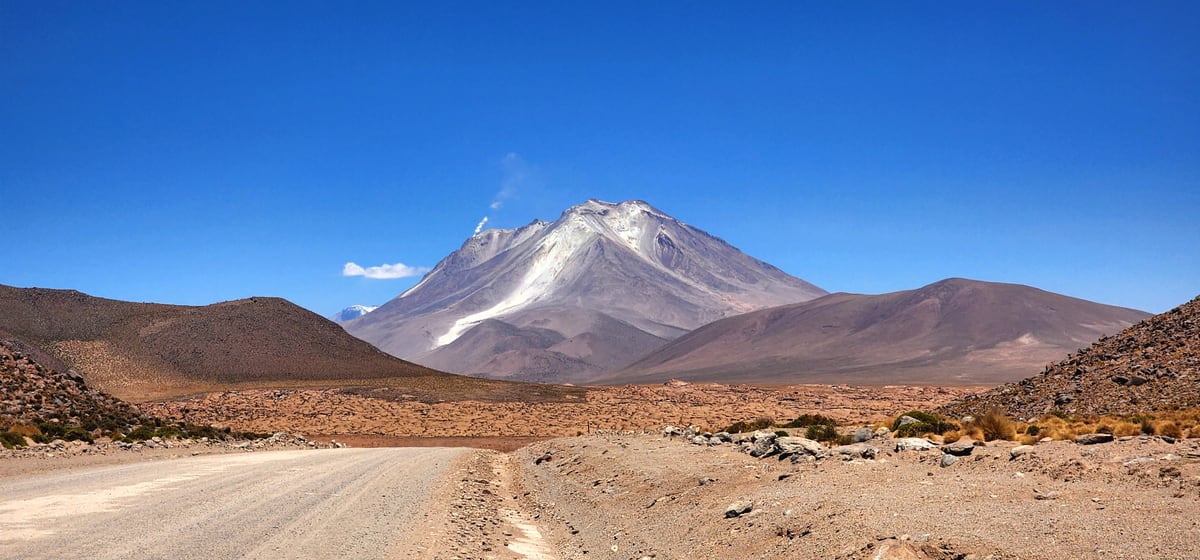 Estrada de terra atravessando o deserto, com um vulcão recortado ao fundo